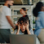 Overwhelmed businesswoman holding her head while working on a laptop in a chaotic office environment with blurred colleagues rushing around