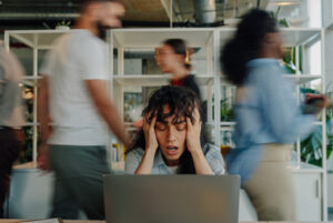 Overwhelmed businesswoman holding her head while working on a laptop in a chaotic office environment with blurred colleagues rushing around
