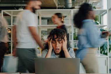 Overwhelmed businesswoman holding her head while working on a laptop in a chaotic office environment with blurred colleagues rushing around